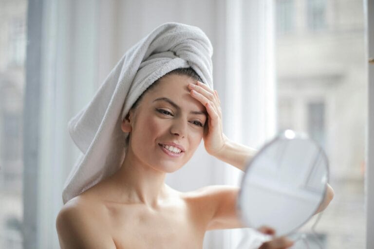 A woman with a towel on her head looking in a handheld mirror and smiling by pexel