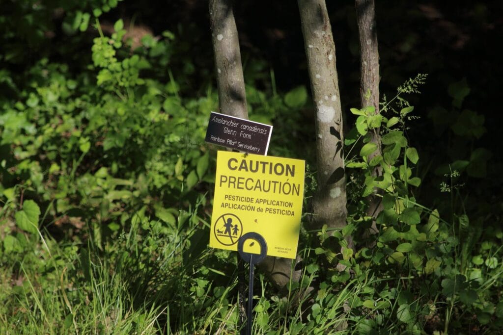 Close-up of a caution sign warning about pesticide application near green plants. uji pestisida.